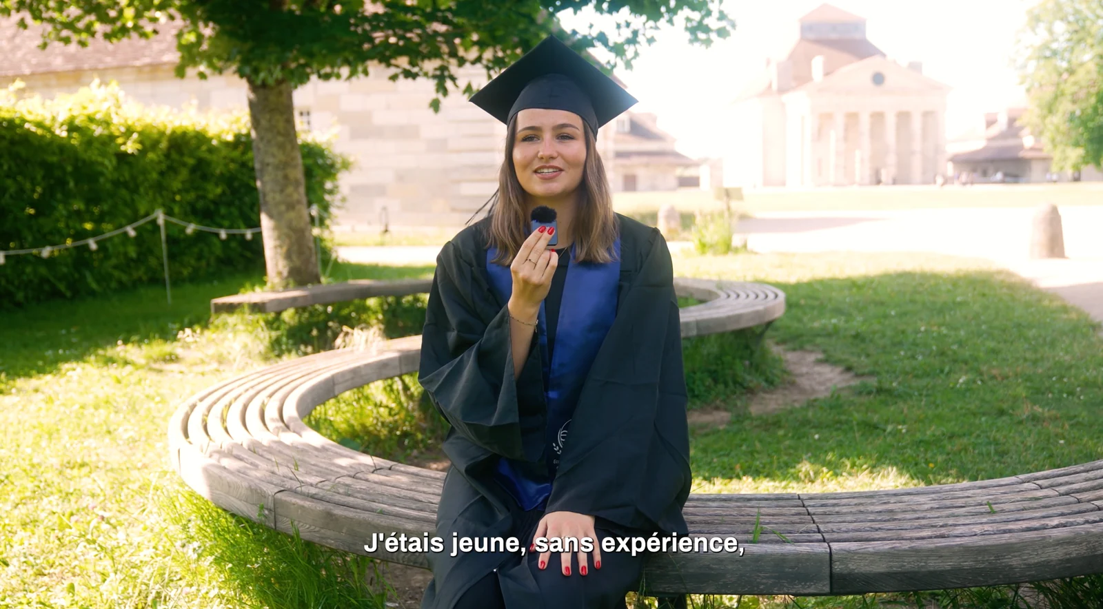 Une personne en tenue de diplômée tient un microphone dans un jardin.