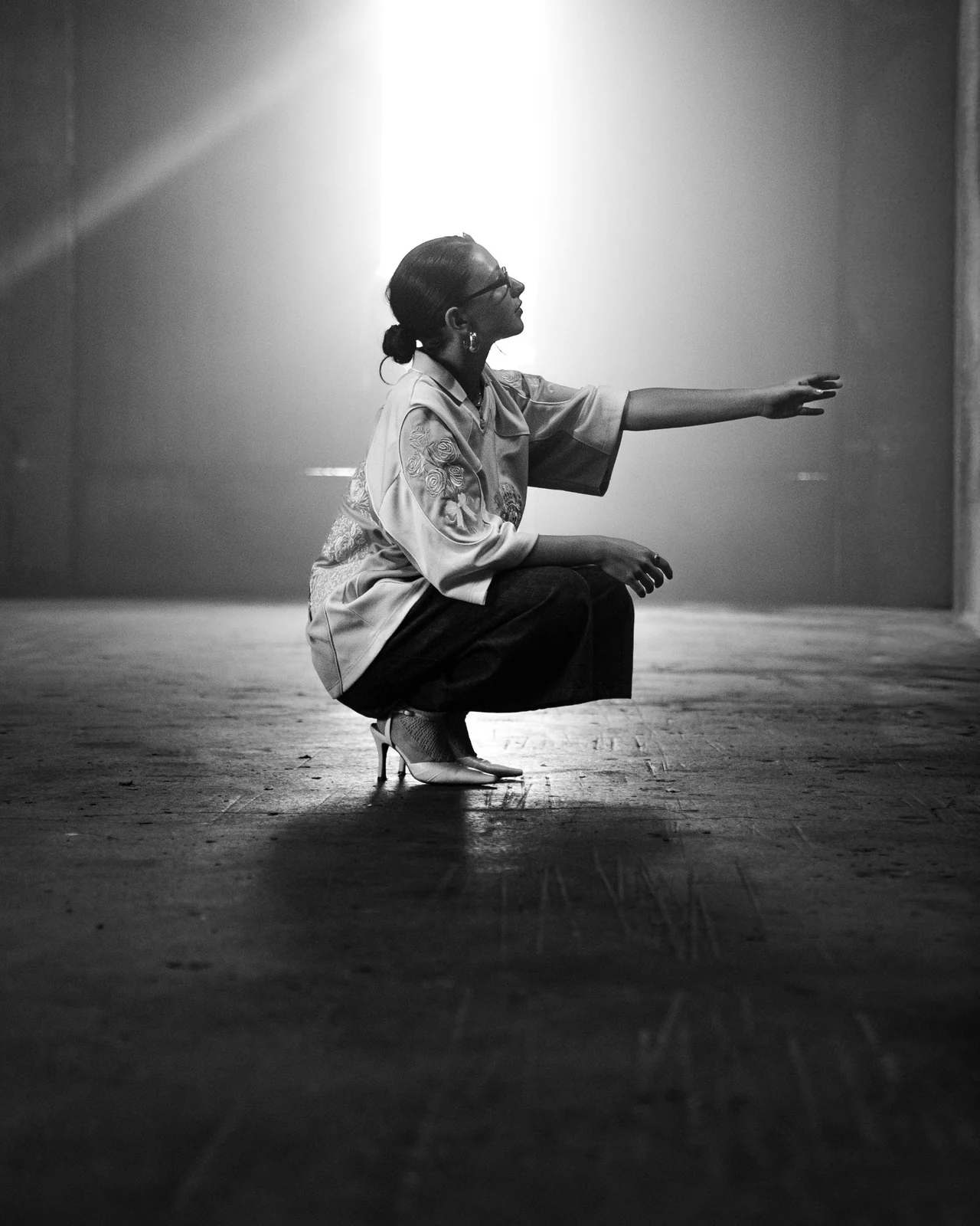 Une personne accroupie, bras tendu dans un faisceau de lumière, en noir et blanc.